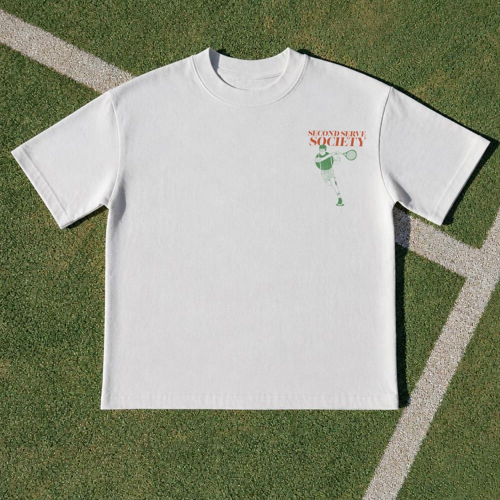Freshly pressed Second Serve Society tee lying flat on a green tennis court, showing the small front chest print